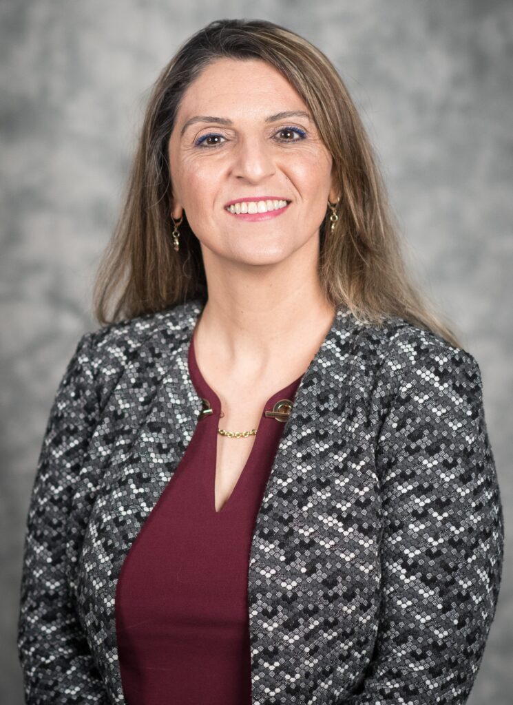 a woman with long, light brown hair, wearing a dark red blouse and a gray patterns jacket smiles warmly at the viewer.